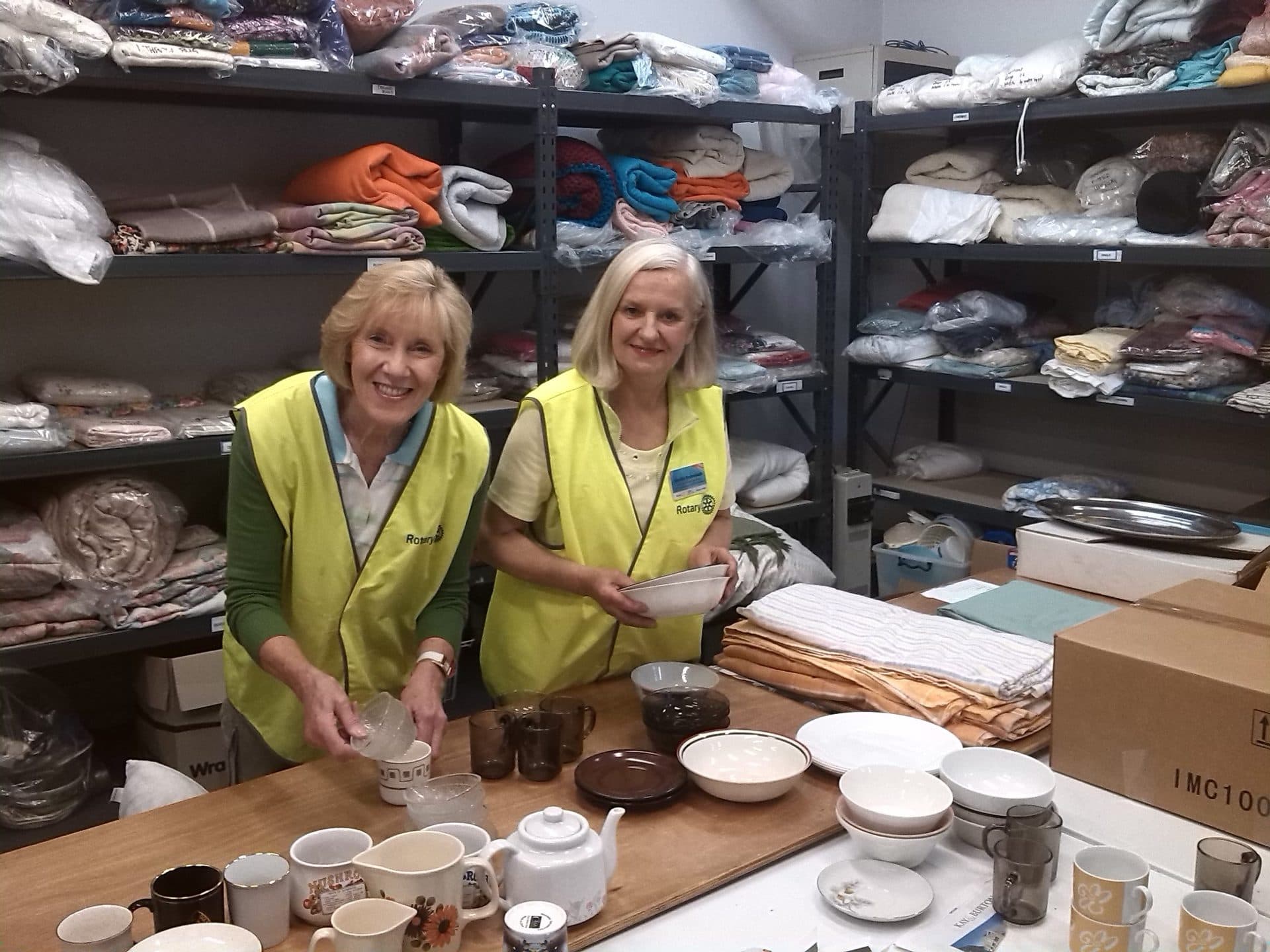 Two WERN volunteers in high-visibility vests packing crockery and kitchenware into starter packs at a workbench.